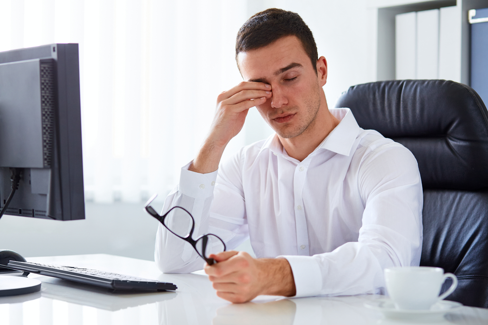 A man holding his glasses rubbing his eye while sitting in front of a computer