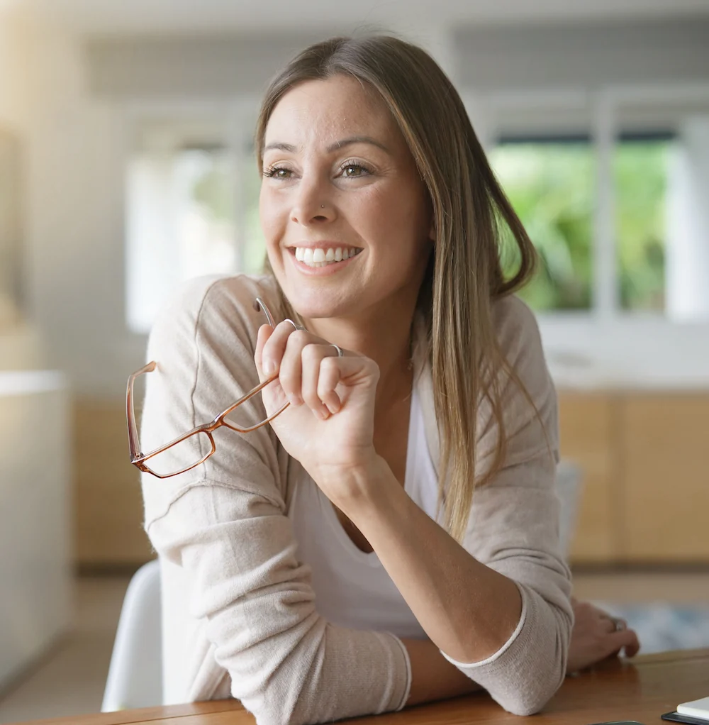 Smiling woman in her 40's sitting at desk holding reading glasses